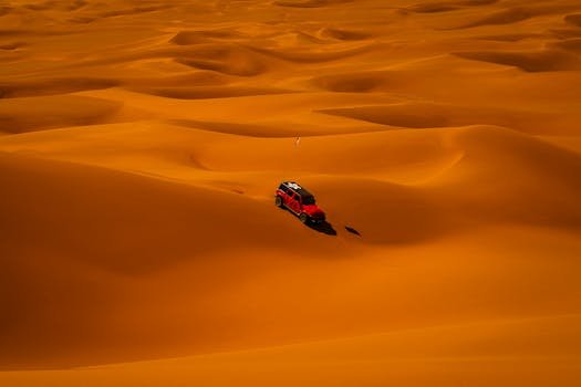 Aerial view of a red off-road vehicle driving through expansive golden desert dunes.